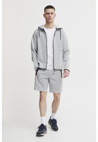 Ensemble de sport gris clair comprenant un sweat à capuche zippé, un T-shirt et un short. Comprend des poches zippées et des accents noirs. Des baskets noires sont portées.