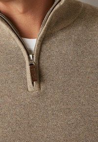 Beige zip-up sweater with a high collar, textured fabric, and a metallic zipper featuring a brown leather pull tab.