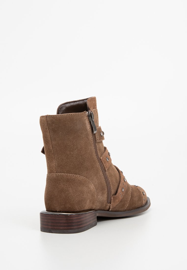 Stivale alla caviglia in suede marrone con zip laterale, borchie metalliche decorative e tacco basso impilato. Il design include diverse cinghie sulla parte anteriore.