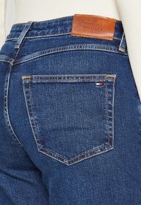 Jeans in denim blu con una vestibilità slim, caratterizzati da un'etichetta in pelle impressa con "TOMMY HILFIGER" e un piccolo dettaglio a bandiera sulla tasca posteriore.