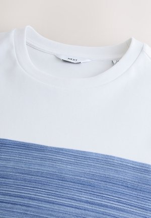 T-shirt blanc avec une bande horizontale texturée en tissu bleu, col rond et matière lisse. Étiquette visible à l'intérieur du col.