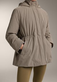 Vrouw draagt een beige en zwart geruit jack met capuchon, handen in zakken en olijfgroene legging tegen een neutrale achtergrond.