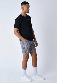 Camiseta negra con logotipo bordado, pantalones cortos estampados en azul, blanco y gris, y zapatillas blancas. El modelo está de pie de lado contra un fondo claro.