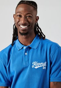 Blauwe polo shirt van gestructureerde stof, met een kraag, twee knopen en een wit geborduurd "Kaporal 04" logo op de linkerborst.