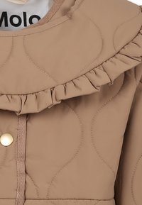 Tan quiltad jacka med volangkrage, guldknäppning och texturerade sömmar. Mjuk, slät tyg med subtil lyster.