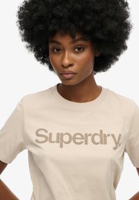 Beige bomulds t-shirt med rund hals og korte ærmer, prydet med et fremtrædende brunt "Superdry" logo på brystet.