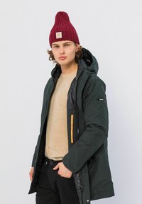 Donkergroene parka jack met een glanzende zwarte voering, oranje ritsdetails en capuchon, gecombineerd met een beige trui en een kastanjebruine gebreide muts.