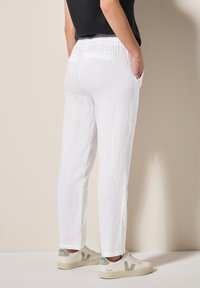 Pantaloni bianchi di lino con taglio dritto, dotati di tasche e una struttura morbida, abbinati a sneakers beige con dettagli verdi.