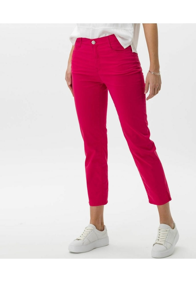 Femme portant un pantalon rose vif longueur cheville, des baskets blanches et un haut blanc rentré, se tenant devant un fond uni.