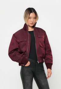 Young woman wearing a maroon bomber jacket with large pockets, black top, and dark textured pants, standing against a plain white background.