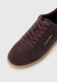 Sneakers en suède marron avec un design à lacets, une semelle en caoutchouc et le logo "Calvin Klein" en blanc. Possède un bout rond et une surface texturée.