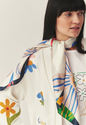 Nainen, jolla on pitkät mustat hiukset, valkoinen neule ja kukkakuvioinen huivi, katsoo oikealle, pukeutunut vaatteisiin ja huiviin, joissa on värikkäitä kukkakuvioita.