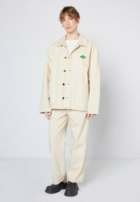 Beige bomuldsjakke med krave, frontknapper, to lommer og et grønt diamantlogo. Båret med matchende beige bukser og sorte sko.
