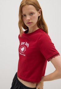 Camiseta corta roja de algodón, con un escote redondo, detalles de encaje en las mangas y un diseño gráfico blanco con detalles florales y de texto.