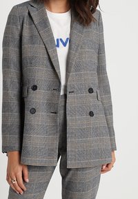 Blazer gris croisé à carreaux, avec des boutons noirs et des revers crantés, porté avec un t-shirt blanc imprimé en dessous.