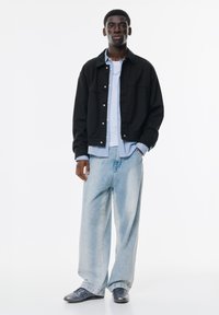 Jachetă neagră oversized, cămașă cu butoni de culoare albastru deschis, blugi largi decolorati, pantofi gri, design casual cu detalii hardware proeminente.