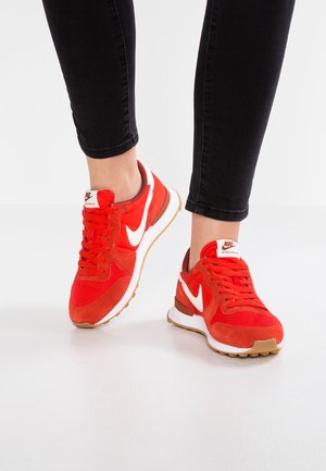 Osoba nosząca jasnoczerwone sneakersy Nike z białymi logo w kształcie znaku Swoosh i czarne dopasowane spodnie, stojąca na białej podłodze.