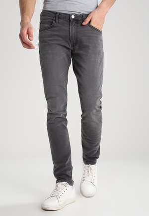 Man wearing slim-fit dark gray jeans, white sneakers, and a light gray shirt with one hand in pocket, walking forward on plain background.