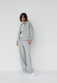 Szara bluza z kapturem na zamek z elastycznymi mankietami oraz pasujące szare spodnie dresowe. Materiał wydaje się miękki, z luźnym krojem. Całość dopełniają białe buty.