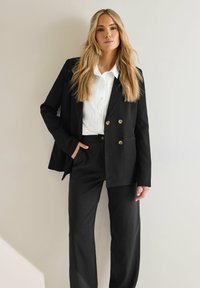 Svart kavaj med dubbelknäppning och mässingsknappar, i kombination med en vit skjorta med krage och svarta skräddarsydda byxor. Slät tygtextur.