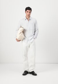 Gestreiftes Hemd in hellblau und weißem Stoff, kombiniert mit lockeren weißen Hosen und schwarzen Schuhen. Das Subjekt hält eine beige Tasche.