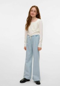 Camicia bianca a maniche lunghe, pantaloni larghi blu chiaro con vita elasticizzata e cordoncino, scarpe nere, modella in piedi contro uno sfondo neutro.