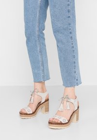 Ljusgrå mocka klacksandaler med blommiga utskärningar, fransar, en staplad träklack och en beige platå. Bärs med avkortade blå jeans.