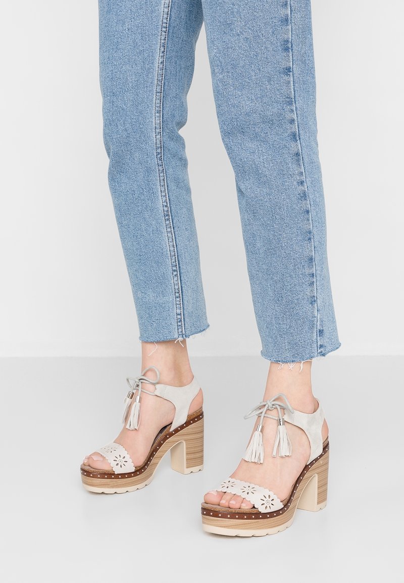 Ljusgrå mocka klacksandaler med blommiga utskärningar, fransar, en staplad träklack och en beige platå. Bärs med avkortade blå jeans.