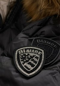 Črn pufast plašč izdelan iz blaga z izstopajočo črno in srebrno okrašeno nalepko z obliko ščita ter besedilom "Blauer." Vezenina je iz žameta.