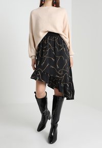 Beige neulepusero, jossa on pudotetut olkaimet, musta hame, jossa on kultainen kuvio ja röyhelöhelma, yhdistettynä kiiltävämustiin polvisukkiin.