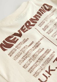 T-shirt beige vintage avec un imprimé brun fané listant les villes du Royaume-Uni et les dates de concerts complets du groupe Nevermind en novembre.