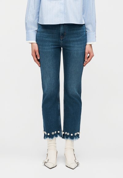 Γυναίκα που φοράει κοντά μπλε τζιν με φθαρμένα τελειώματα διακοσμημένα με πέρλες και λουλουδένιες χάντρες, συνδυασμένα με λευκές κάλτσες και μυτερά παπούτσια.