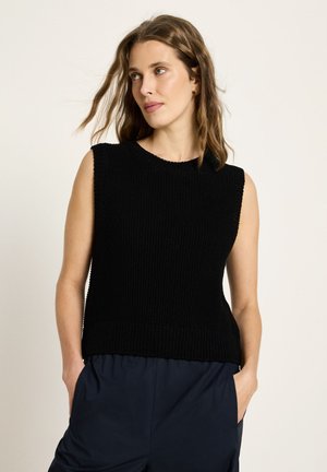 Frau mit schulterlangem braunem Haar, die ein ärmelloses schwarzes Strickoberteil und dunkle Hose trägt, die Hände in den Taschen, Blick zur Seite gerichtet.