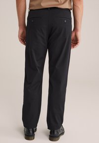 Vista posteriore di una persona che indossa pantaloni neri con tasche posteriori con bottone e scarpe in pelle nera, in piedi su uno sfondo chiaro.