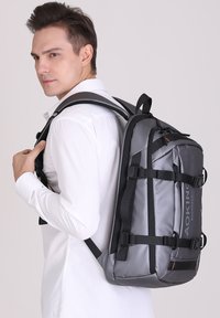 Zaino grigio con spallacci neri, dal design elegante, dotato di fibbie regolabili e dettaglio del logo. Tessuto strutturato con forma rigida.