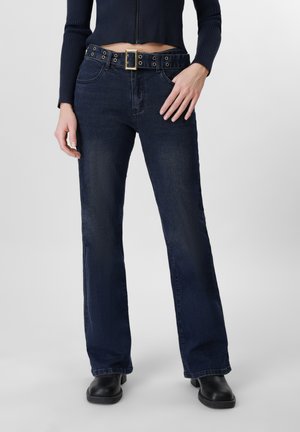 Person iført mørkeblå bootcut-jeans med et bredt sort bælte med metaløjer og sorte ankelstøvler, stående mod en ensfarvet baggrund.