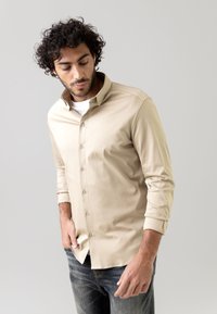 Vyras su garbanais plaukais, vilkintis smėlio spalvos marškinį su ilgomis rankovėmis ir mygtukais ant baltos marškinėlių, bei tamsias džinsus, žiūri žemyn, viena ranka kišenėje.