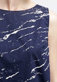 Námořnická modrá bezrukávová halenka s abstraktním bílým vlnkovým vzorem, vyrobená z hladké látky. Má kulatý výstřih a čisté okraje.