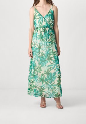 Kobieta nosząca sukienkę maxi bez rękawów z dekoltem w serek, w zielono-niebieski wzór liści, z talią wciętą i sandałami z przezroczystymi paskami.