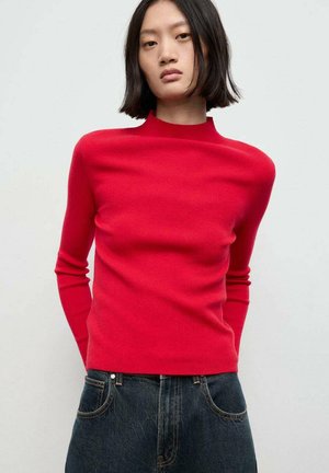 Giovane donna con capelli neri lisci che indossa un top rosso a maniche lunghe aderente e jeans blu scuro, in piedi contro uno sfondo chiaro e neutro.