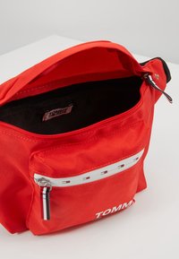 Rode nylon schoudertas met een ritsvak en "TOMMY" branding. Heeft een gestructureerd oppervlak en een contrasterende zwarte binnenkant. Verstelbare schouderriem.