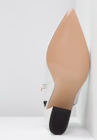 Zapato de tacón alto blanco con punta afilada, suela texturizada beige, base del tacón negra y un detalle decorativo en la tira lateral.