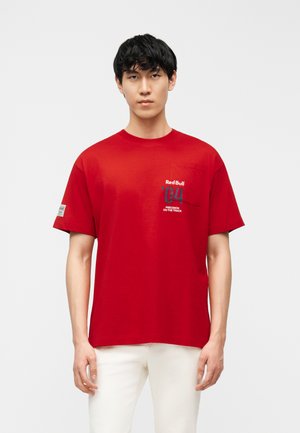 Άνδρας που φοράει κόκκινη κοντομάνικη μπλούζα Red Bull '04 με τσέπη στο στήθος και λευκό παντελόνι, στέκεται μπροστά σε απλό φωτεινό φόντο.