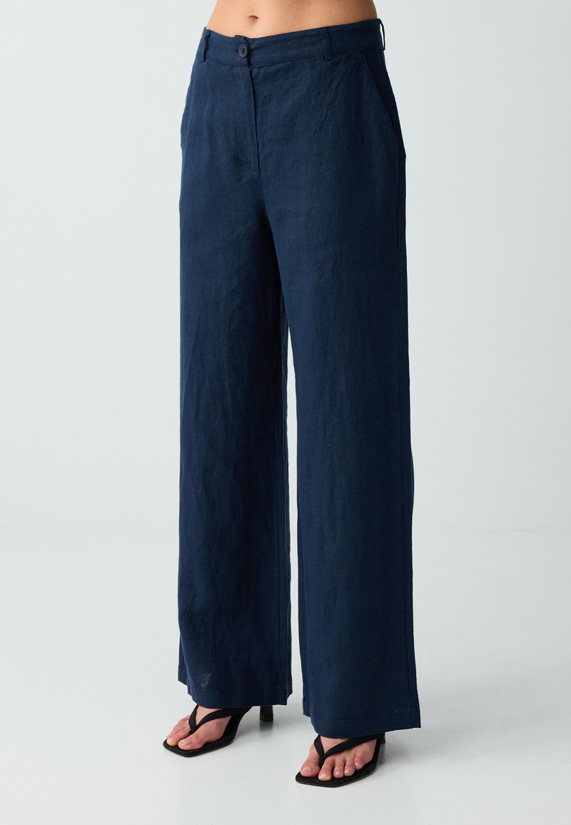 Femme portant un pantalon large taille haute bleu marine et des sandales à talons noires à fines brides sur un fond clair uni.