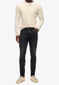 Camisa henley de manga larga en beige claro con tres botones, combinada con jeans de mezclilla oscura y zapatillas crema, accesorios con un cinturón negro.