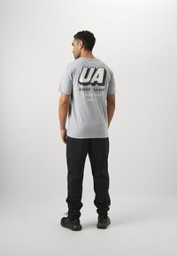 Grijze T-shirt met korte mouwen met zwarte grafische tekst "UA BODY SHOP" en "Quality Service Since 96" op de achterkant, gecombineerd met zwarte broeken en schoenen.