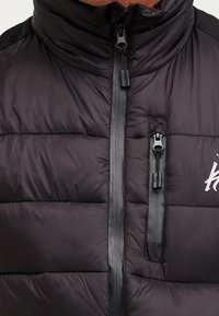 Chaleco de plumas negro con un alto cuello, diseño acolchado horizontal, cremallera frontal y detalle del logo en blanco en el lado izquierdo.