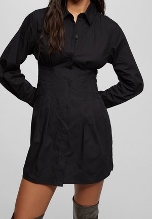 Woman wearing a fitted black button-up mini dress with long sleeves and a collar, paired with knee-high black boots.