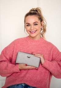 Gray wallet with embossed dot pattern and black zipper; held against a pink knit sweater. Logo in the center of the wallet.