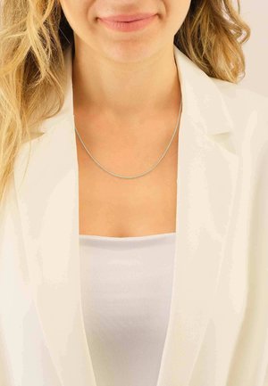 Collier en chaîne argentée porté sur un top blanc, avec une texture lisse et un design délicat, complété par un blazer de couleur claire.
